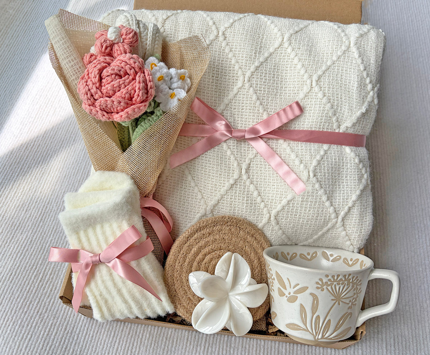 A gift box containing a cream-colored diamond-knit blanket tied with a pink ribbon, a pink crocheted flower bouquet wrapped in burlap, folded cream socks with pink ribbon bows, a round woven coaster, a white flower-shaped object, and a cream ceramic mug with a floral pattern.