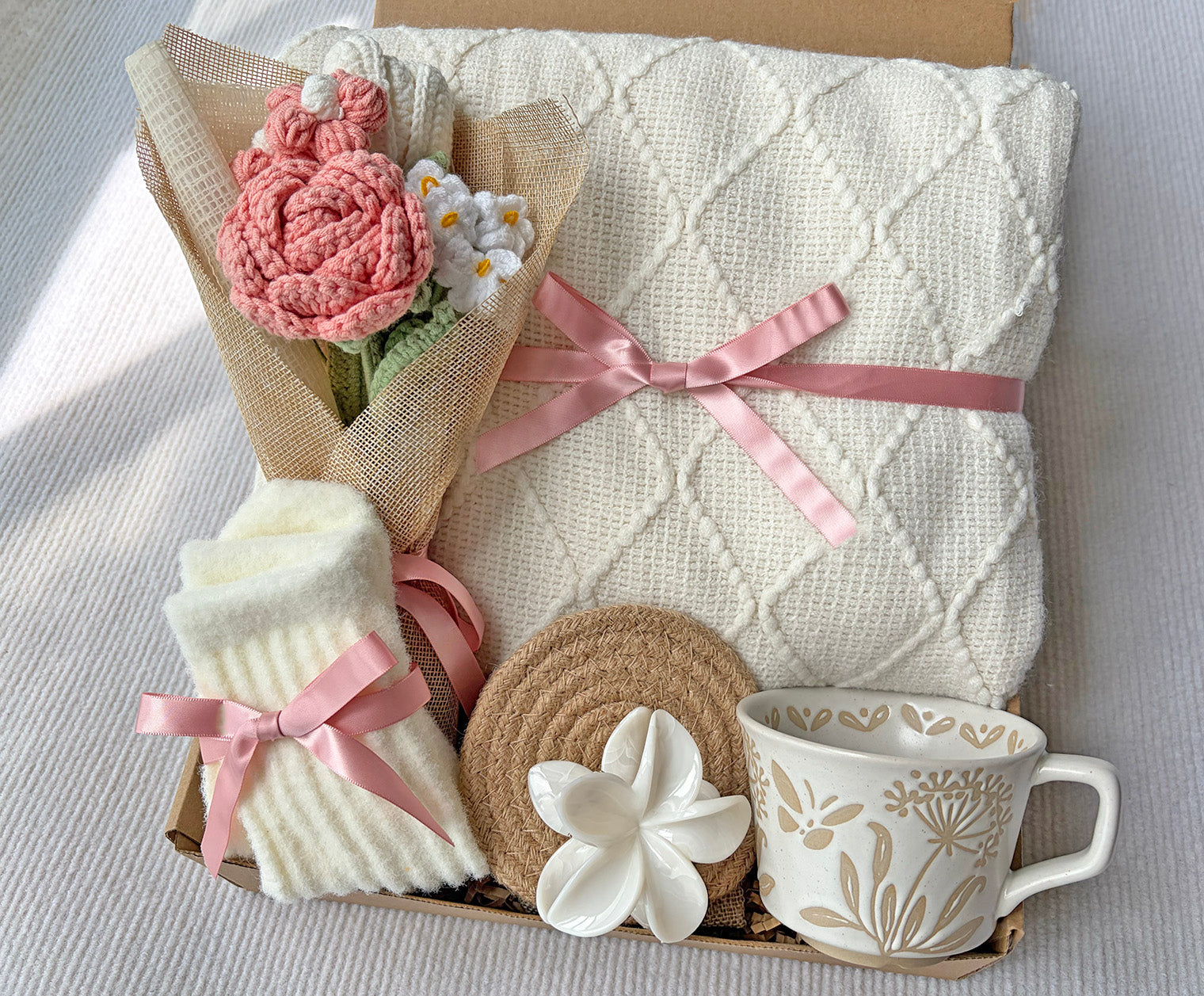 A gift box containing a cream-colored diamond-knit blanket tied with a pink ribbon, a pink crocheted flower bouquet wrapped in burlap, folded cream socks with pink ribbon bows, a round woven coaster, a white flower-shaped object, and a cream ceramic mug with a floral pattern.